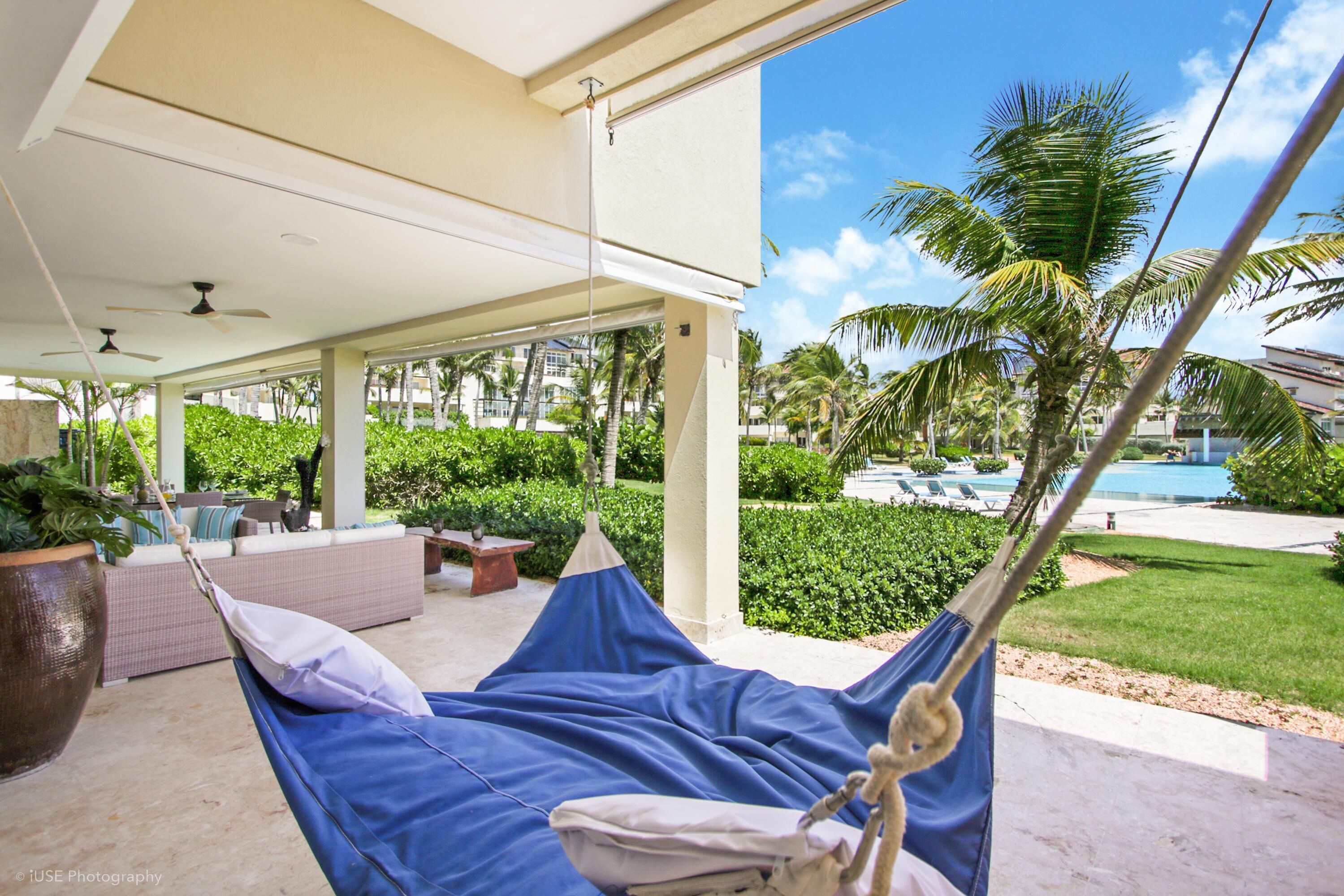 A hammock under a covered patio with a view of a pool and palm trees.