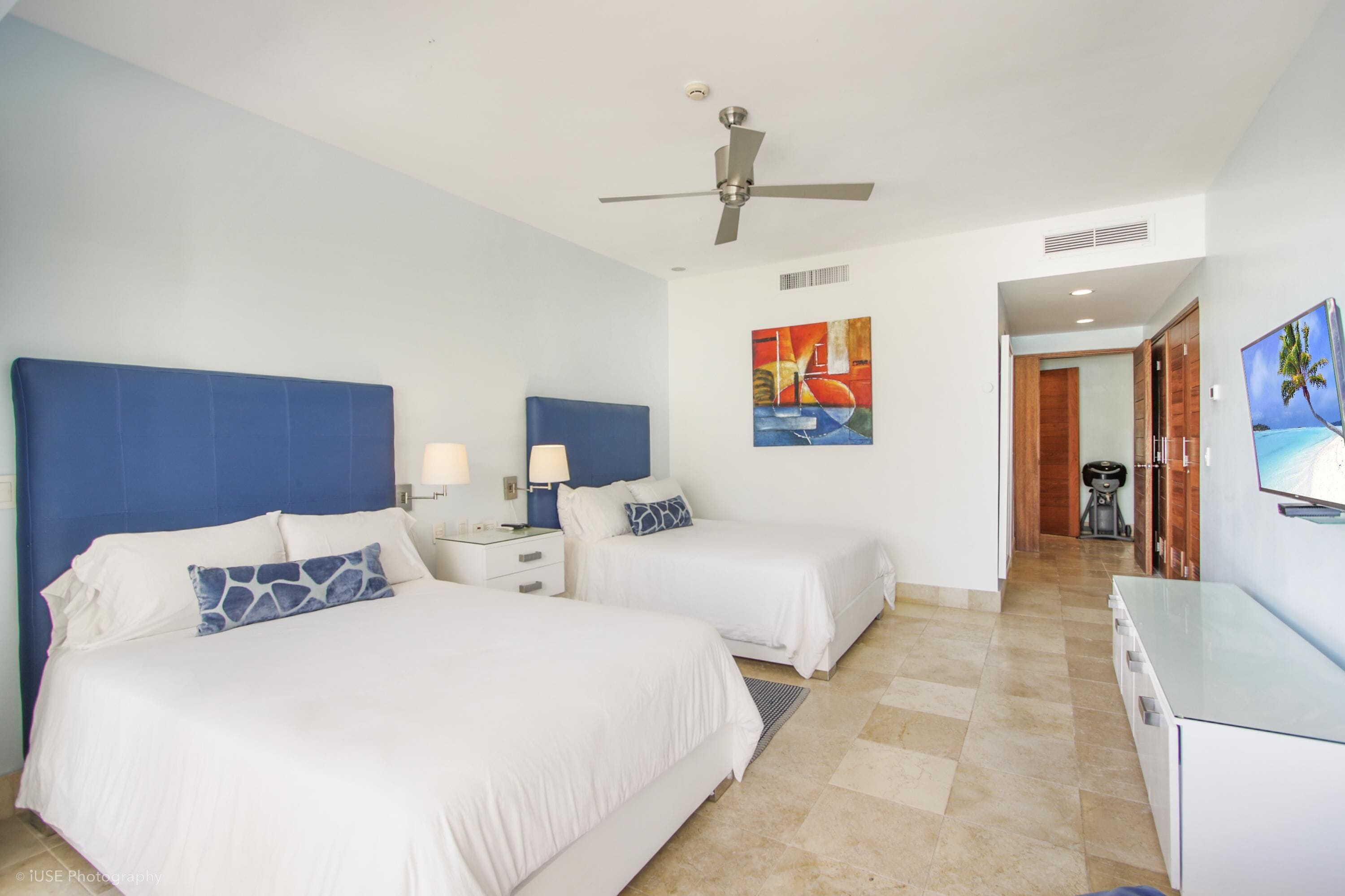 A hotel room with two beds, a ceiling fan, artwork on the walls, and a TV on the wall.
