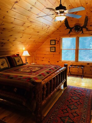 A cozy attic bedroom with a wooden ceiling, a large bed with a colorful quilt, a standing lamp, and a window with a view. - Log Home Nestled Between The White Mountains and The Lakes Region (Campton)