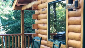 A log cabin with a wooden deck, two rocking chairs, and a window with a view of the surrounding forest.