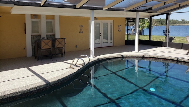 A covered swimming pool area with a view of a lake, a patio with a table and chairs, and a door leading to a covered entrance.