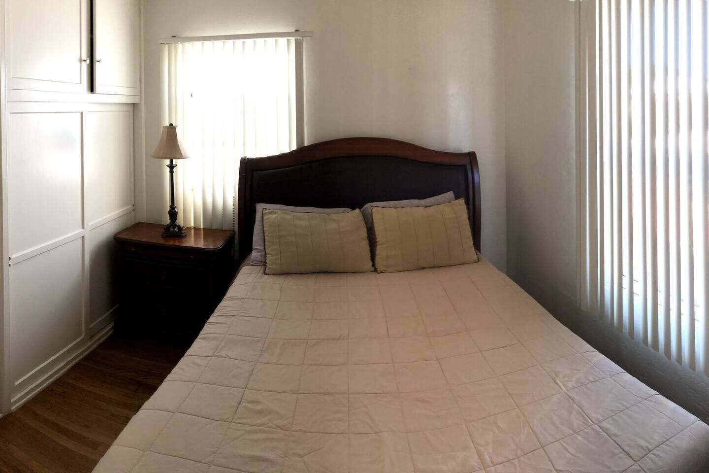 A bedroom with a large bed, two pillows, a nightstand with a lamp, and a window with blinds.