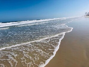 Une plage où les vagues viennent doucement lécher le rivage, un ciel dégagé et des bâtiments qui se dessinent à l’horizon.