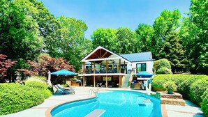Una piscina con trampolín, rodeada por un deck de ladrillo y una terraza de madera con sillas reclinables.