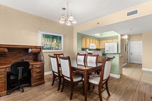 A dining area with a wooden table and chairs, a kitchen in the background, and a painting on the wall. - Ariel Dunes I 1409*Extra bunk room and 3 pools! (Miramar Beach)