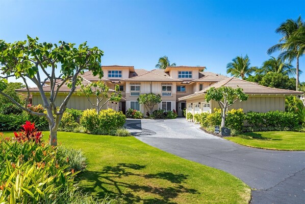 A multi-story residential building with a driveway leading to the entrance, surrounded by well-maintained lawns and tropical plants.