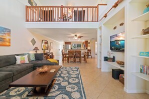 A spacious living area with a dark brown sofa, a coffee table, and a patterned rug. A dining area with wooden chairs and a table is visible in the background. - Hilton Pool Pass  for stays in 2026. Waikoloa Beach Villas C23 (Waikoloa Beach Resort)