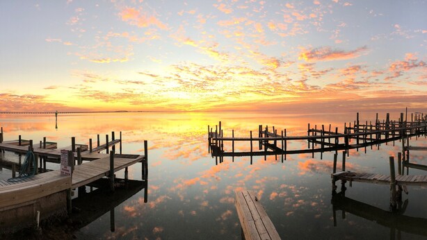 Une jetée au bord de l’eau, avec des passerelles et des quais en bois qui s’étendent dans des eaux calmes, reflétant le ciel au coucher du soleil.