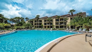 A swimming pool surrounded by lounge chairs and palm trees, with a multi-story building in the background.