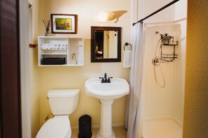 A bathroom with a toilet, pedestal sink, and shower. There is a framed picture above the sink and a towel rack on the wall.