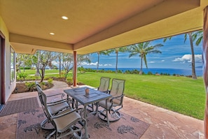 A covered outdoor seating area with a table and chairs, overlooking a well-maintained lawn and palm trees, with a view of the ocean.