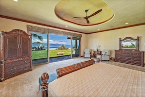 Quarto espaçoso com uma cama grande, ventilador de teto e vista para uma área gramada e oceano através de amplas portas de correr.