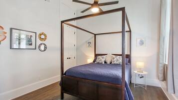 Een hemelbed met een donker houten frame, een blauwe spreidek en gekleurde kussens. Er is een klein nachtkastje met een lamp, een spiegel aan de muur en kunstwerken in lijsten. De kamer heeft houten vloeren en een plafondventilator.