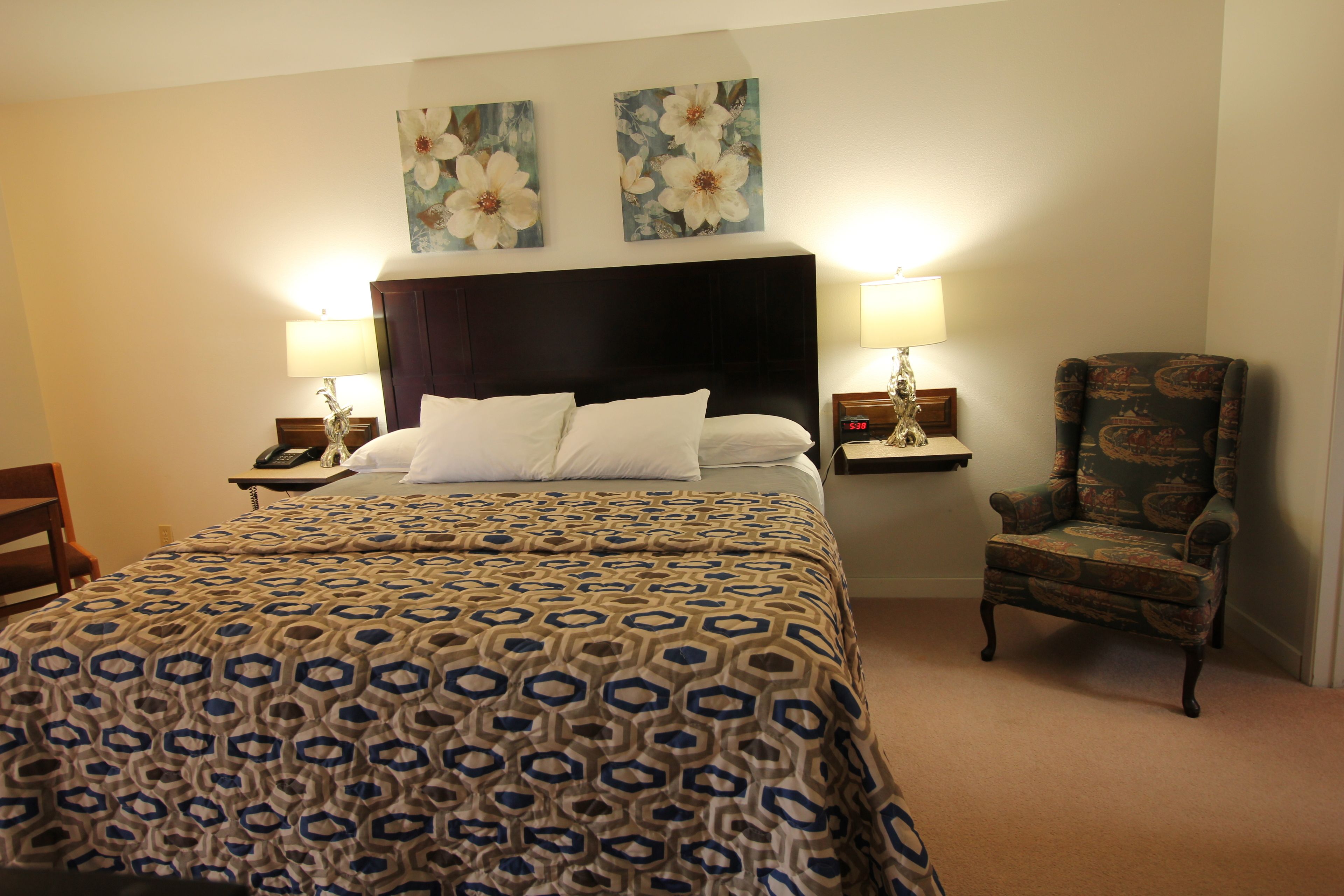A hotel room with a large bed, two wall-mounted lamps, a chair, and two floral paintings.