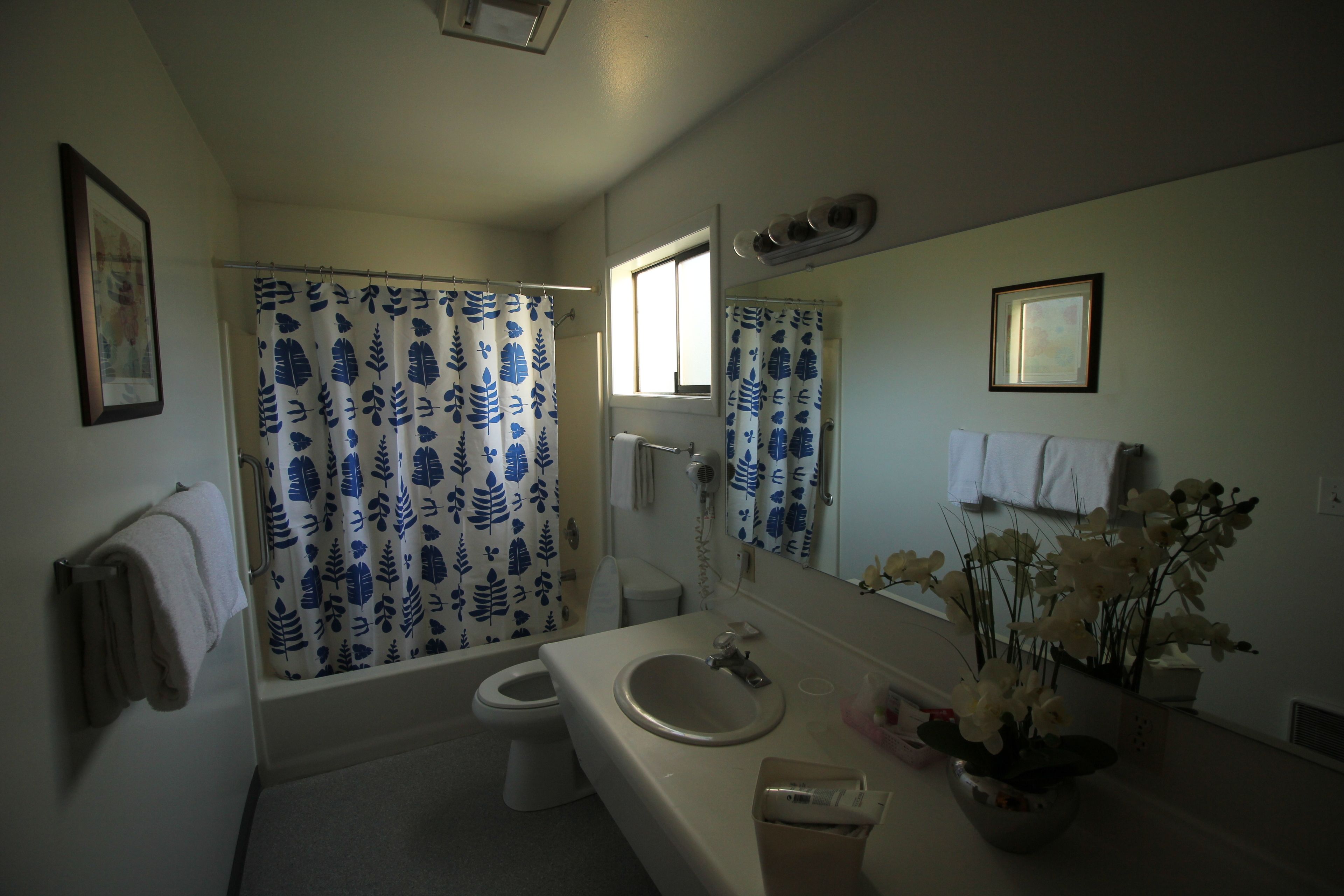 Un baño con bañera, inodoro y lavamanos. Hay una cortina de ducha con estampado de hojas, un espejo y un florero con flores sobre el lavamanos.