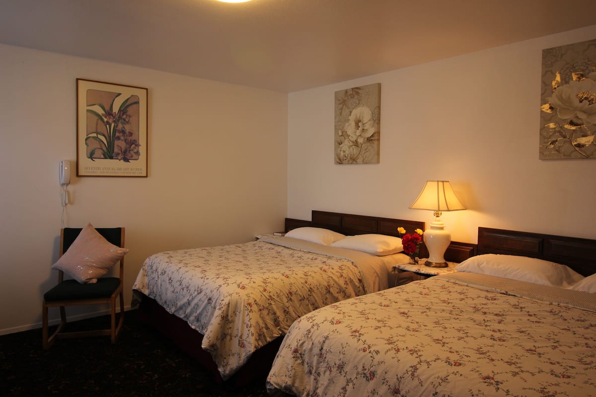 A hotel room with two beds, a chair, a lamp, and floral artwork on the walls.