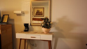 Un petit bureau blanc avec un seul tiroir, une machine à café et une plante en pot. Un tableau encadré est accroché au mur, au-dessus du bureau.