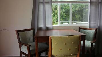 A dining area with a wooden table and two upholstered chairs, a window with a view of greenery, and a curtain partially drawn.