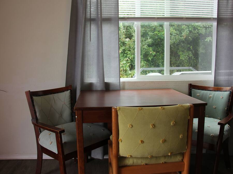 A dining area with a wooden table and two upholstered chairs, a window with a view of greenery, and a curtain partially drawn.