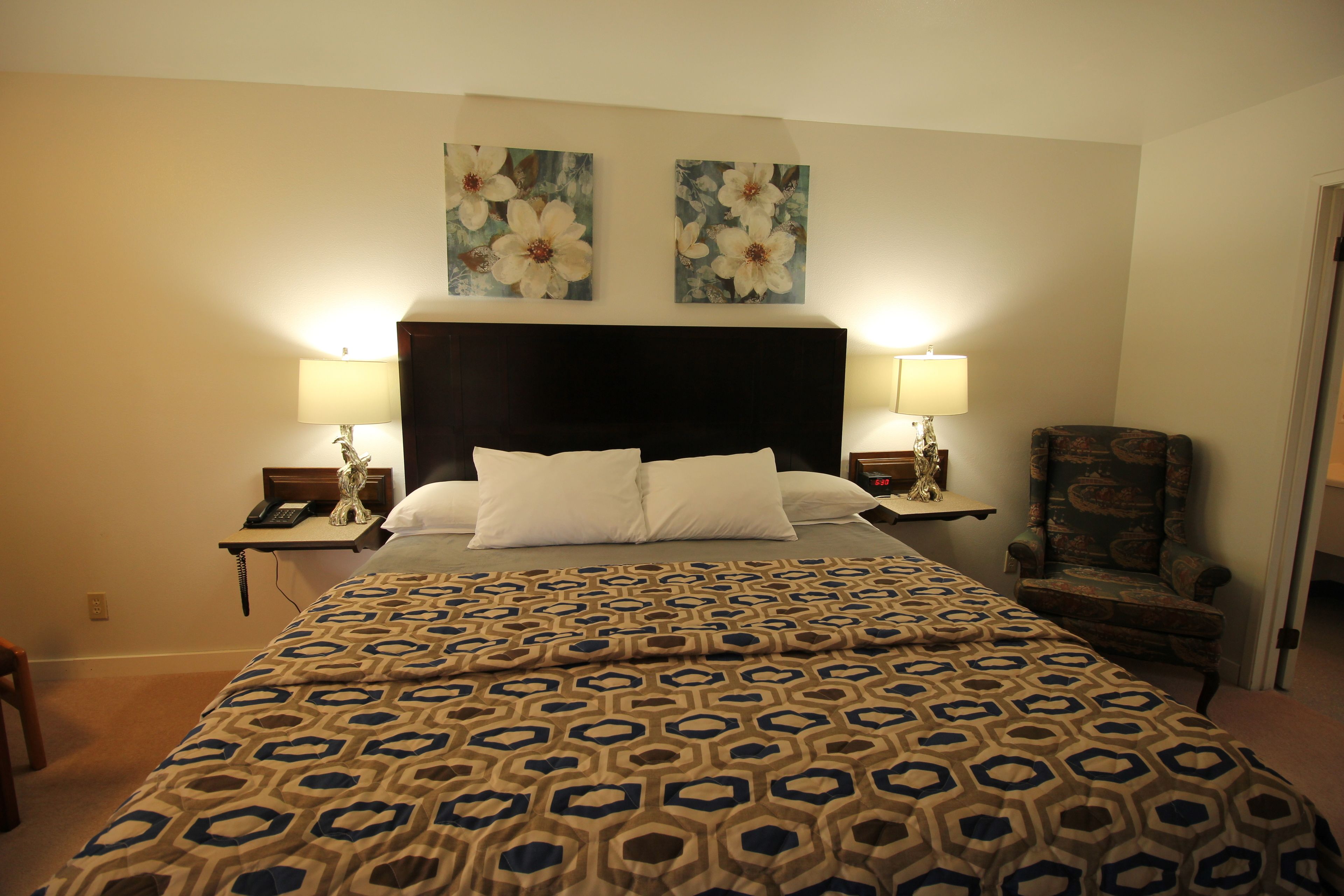 A hotel room with a large bed, two wall-mounted lamps, a chair, and two floral paintings on the wall.
