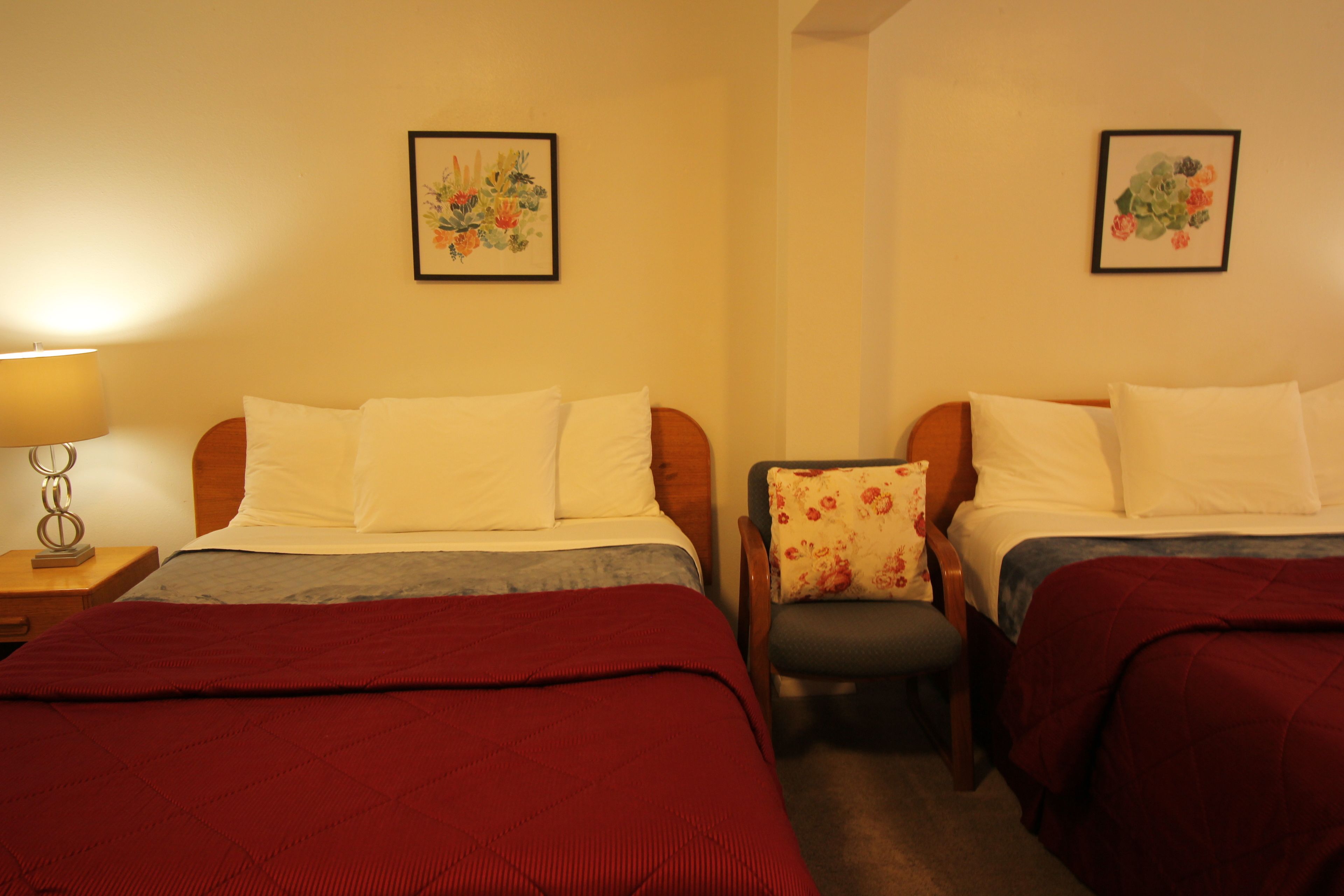 A hotel room with two beds, each with white bedding and a red blanket. There is a chair with a floral cushion, and two framed floral paintings on the walls.