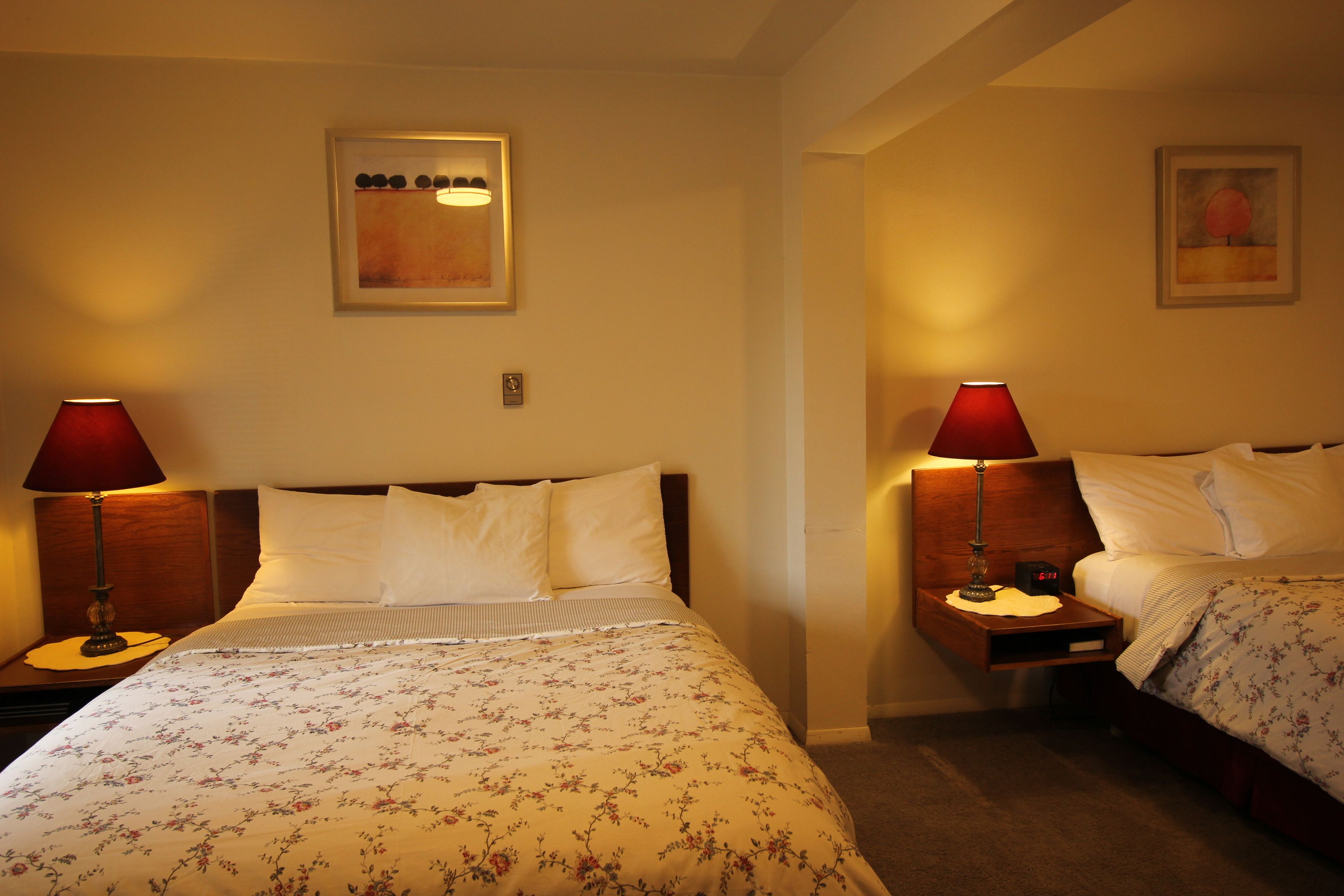 A hotel room with two beds, each with a wooden headboard, white bedding, and a red table lamp. There are framed artworks on the walls and a small clock on the bedside tables.