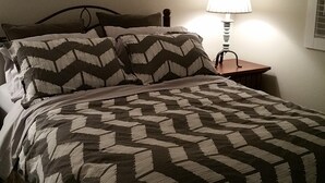 A neatly made bed with a patterned bedspread and two pillows, a bedside table with a lamp, and a wall-mounted air conditioning unit. - New Construction Coach House, 10 Minute Walk to Downtown and Ferry Spotless (Nanaimo)