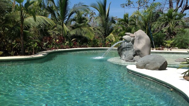 Ein klares Schwimmbecken mit Wasserfall, umgeben von tropischer Vegetation.