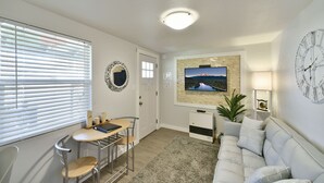 Ein Wohnzimmer mit einer Couch, einem an der Wand befestigten Fernseher, einer Deckenleuchte, einer Uhr und einem Fenster mit Jalousien.