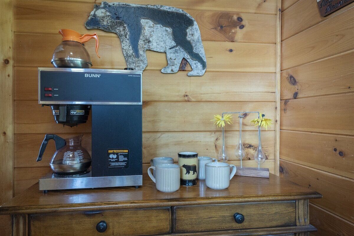 A wooden cabin interior with a Bunn coffee maker, a bear-themed mug, and a small vase with flowers.