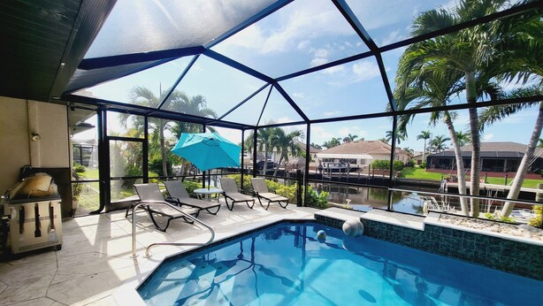 Zona de piscina con sillas de descanso y una sombrilla azul, rodeada por un cerramiento de cristal con vistas a palmeras y casas.