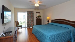 A hotel room with a bed, a television, a desk with a chair, a ceiling fan, and a window with curtains.