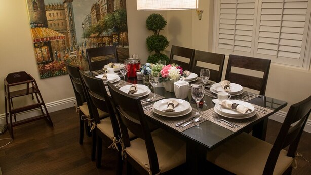A dining table set for six with chairs, a painting on the wall, a small plant, and a window with blinds.