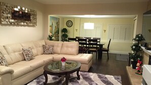 A living room with a beige sectional sofa, a glass coffee table, and a striped rug. There is a dining area with a table and chairs, and a Christmas tree.