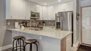 Cozinha moderna com armários brancos, geladeira de aço inoxidável, micro-ondas e painel de azulejos. Há duas banquetas na ilha.