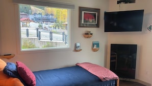 Een kamer met een bed, een raam met uitzicht op gebouwen en bomen, een televisie en wandplanken met foto's.