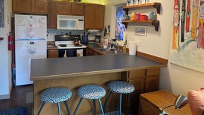 Een keuken met houten kastjes, een witte koelkast, een fornuis, een magnetron en een spoelbak. Er staan drie barkrukken bij het keukeneiland.