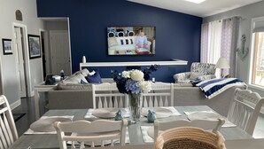 Un salon moderne avec un mur d’accent bleu, un coin repas avec des meubles blancs et un téléviseur fixé au-dessus du canapé.