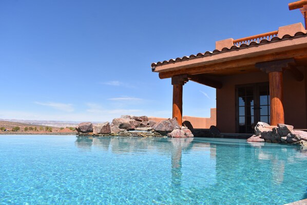 Una piscina azul cristalina con rocas, una pérgola de madera y vistas al paisaje desértico.