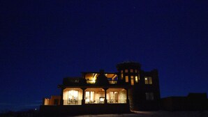 Une grande maison à plusieurs étages, dotée d’un balcon et d’une structure en forme de tour, illuminée la nuit.