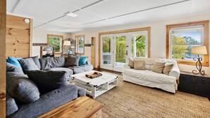 Un salón espacioso con un sofá gris, un sofá blanco y una mesa de centro de madera. Hay ventanas amplias con vistas a zonas verdes.