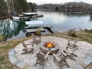 Un espace extérieur circulaire aménagé avec des sièges, doté d’un foyer, entouré de fauteuils Adirondack, et offrant une vue sur un lac où sont amarrés des bateaux, avec des maisons en arrière-plan.