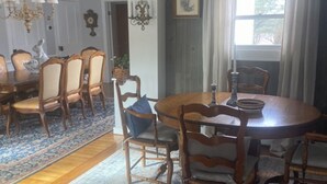 A dining room with a round wooden table, six wooden chairs, a chandelier, and a large window with curtains. - Windham/Hunter All-season French Country Farmhouse-Moments Away from Hayfield. (Maplecrest)