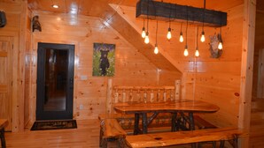 Un intérieur en bois avec une grande table à manger et des bancs, un escalier et une porte donnant sur une autre pièce.