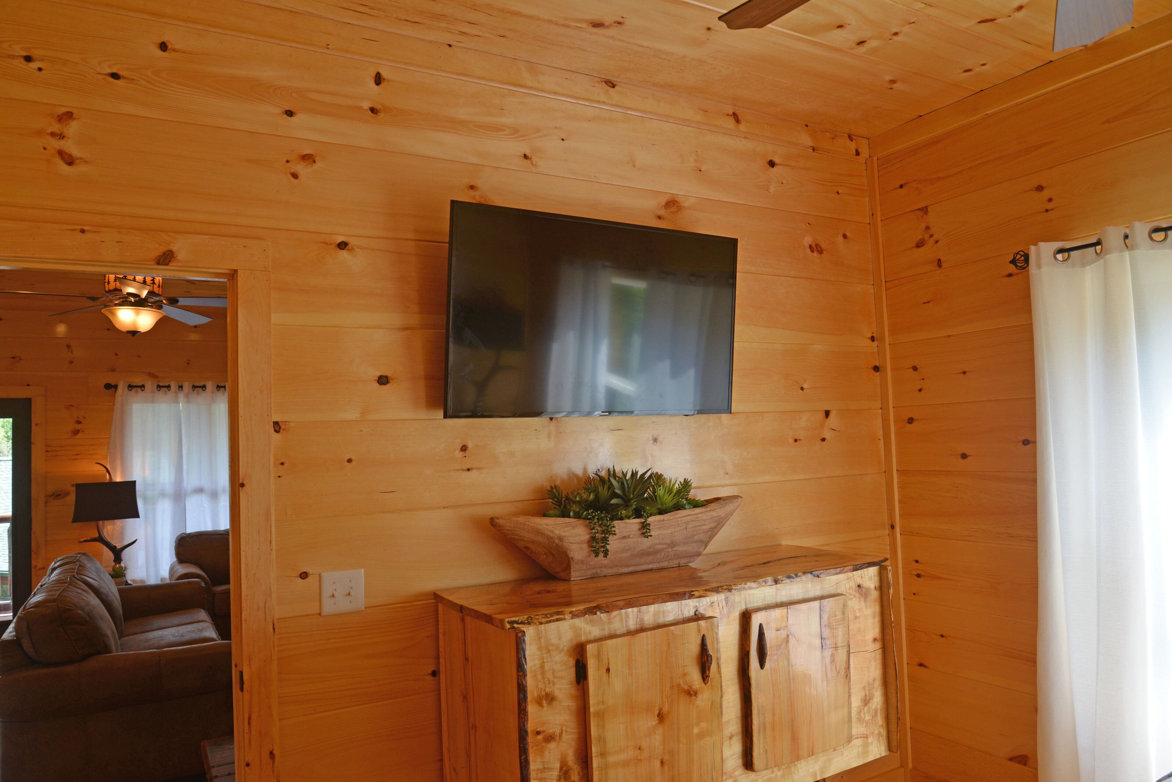 Interieur van een houten chalet met een plafondventilator, een wandgemonteerde flatscreen-tv, een houten kastje met een plant en een bank.