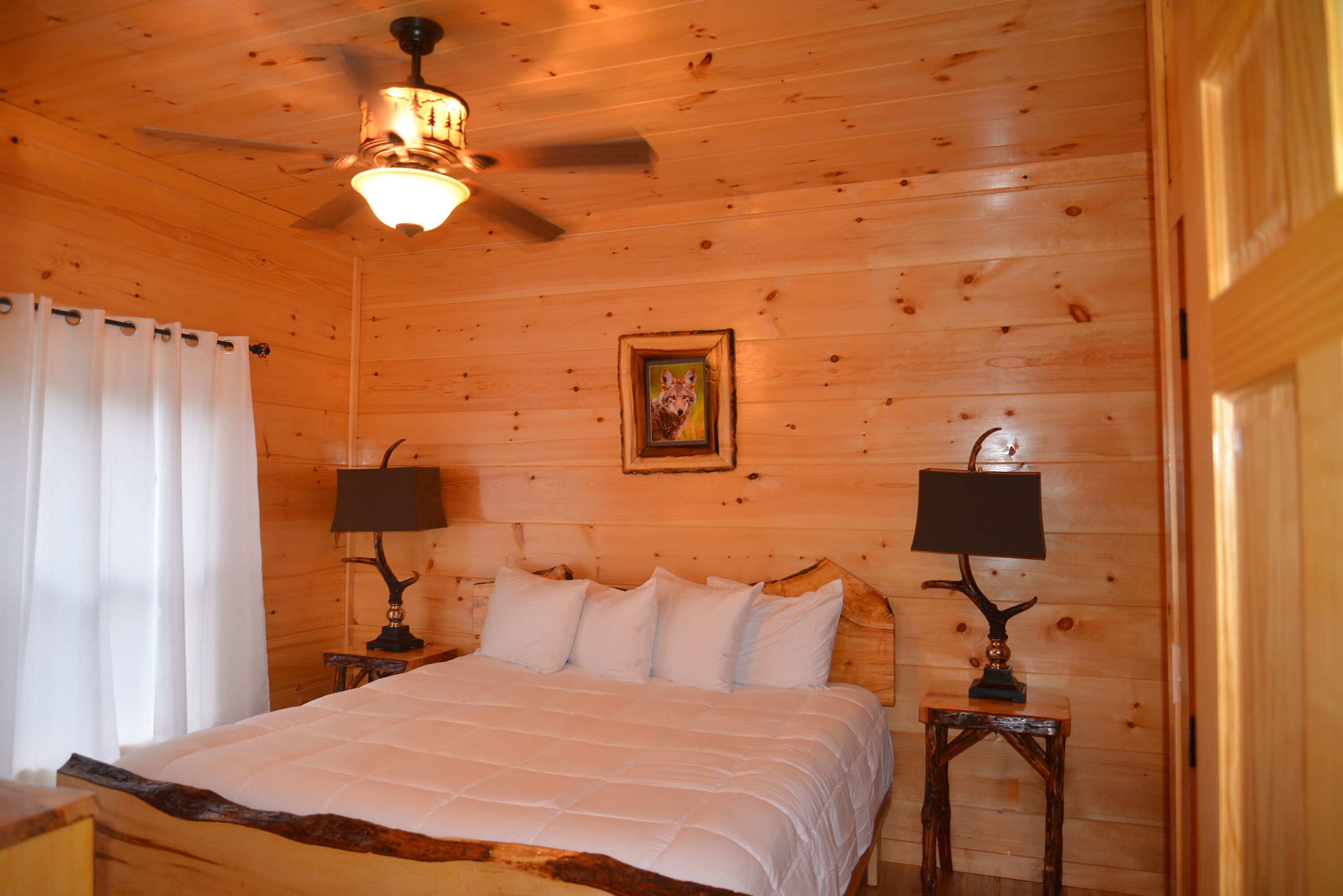 Un dormitorio de cabaña de madera con una cama, dos mesitas de noche con lámparas, un ventilador de techo y un cuadro en la pared.