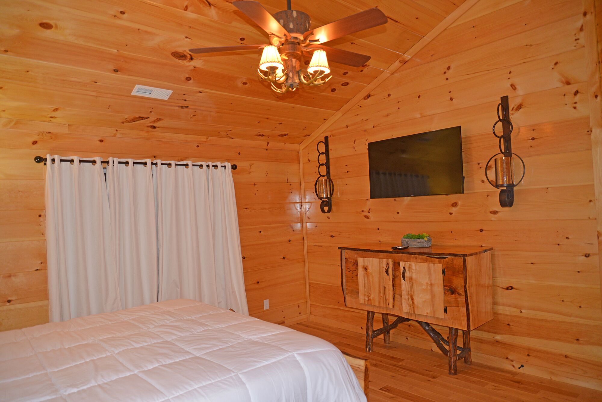 Ein Zimmer einer Holzhütte mit einem Bett, einem Deckenventilator, einem kleinen Tisch und einer Wandleuchte.