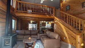 Interior acogedor de una cabaña con una escalera de madera, una sala de estar con sofá y mesa de centro, y una cocina visible a través de una puerta abierta.
