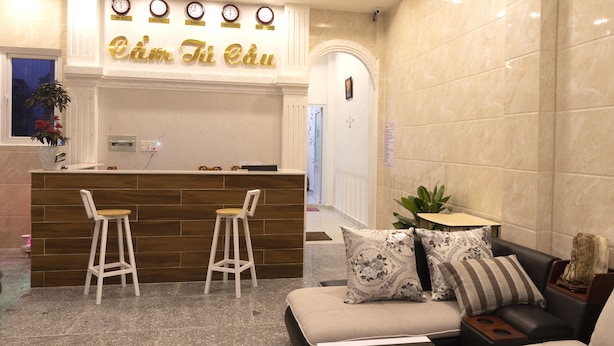 A reception area with a wooden counter, two bar stools, a sofa with patterned cushions, and a clock above the counter displaying times for New York, Sydney, Paris, and Singapore. - MrLee Dalathouse (Xuan Huong - Da Lat)