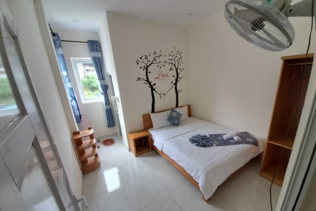 Un dormitorio con cama, mesita de noche, ventilador de pared, ventana con cortinas y una escalera que sube a un piso superior.